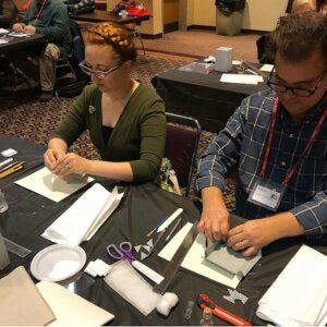 Two people working with paper, scissors and glue to make folded enclosures